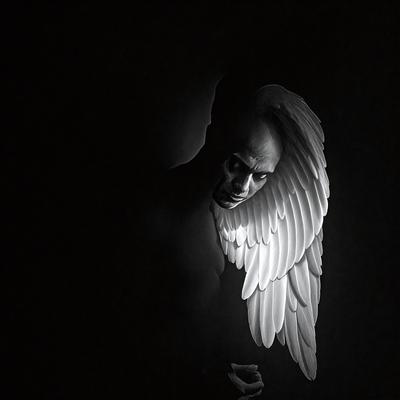 The primary subject of the image is a person with a white angel wing on their back. The setting is a dark room, and the visual style appears to be a combination of a photo and digital art. The dominant colors are black and white, creating a moody atmosphere.